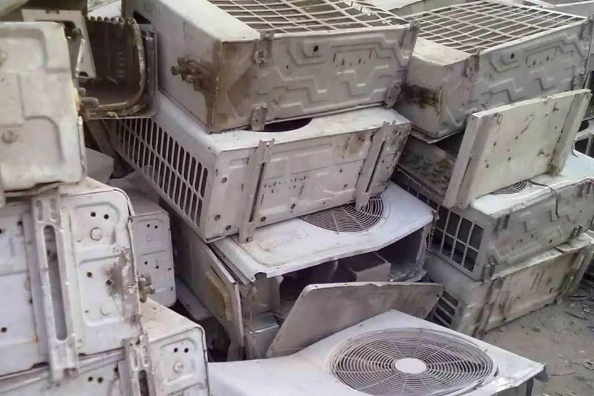 Scrap yard collection of old outdoor air conditioner units and refrigerators stacked for metal recovery and safe gas dismantling.